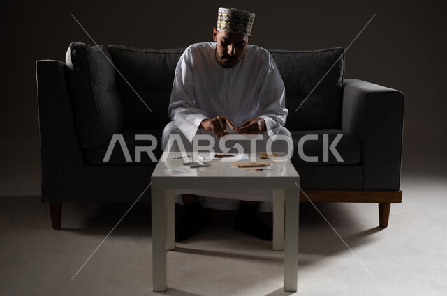 القيام بعادات سلوكية ضارة وتعاطي المواد السامة، الأثار السلبية للادمان على التبغ والمخدرات، المعاناة من الاضطرابات النفسية والمشاكل الصحية، بورتريه لرجل عربي خليجي عماني يرتدي الدشداشه والكمه يجلس على اريكة ويقوم بلف سجائر الحشيش المخدر، صوره ظليه