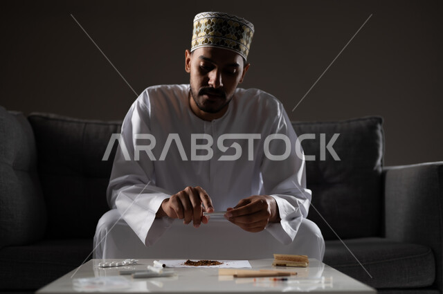 القيام بعادات سلوكية ضارة وتعاطي المواد السامة، الأثار السلبية للادمان على التبغ والمخدرات، المعاناة من الاضطرابات النفسية والمشاكل الصحية، بورتريه لرجل عربي خليجي عماني يرتدي الدشداشه والكمه يجلس على اريكة ويقوم بلف سجائر الحشيش المخدر، صوره ظليه