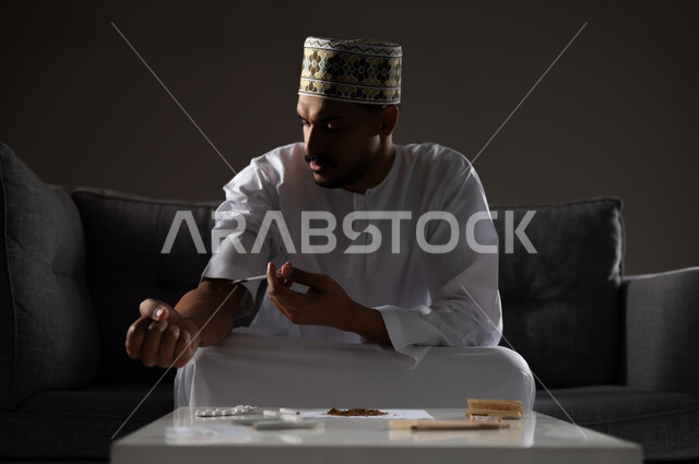 المعاناة من الاضطرابات النفسية والمشاكل الصحية، الآثار السلبية للادمان، القيام بعادات سلوكية ضارة وتعاطي المواد السامة، بورتريه لرجل عربي خليجي عماني يرتدي الدشداشه والكمه يجلس على الاريكة ويقوم بحقن المواد المخدره في اورده الذراع، صوره ظليه