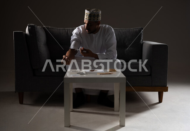 المعاناة من الاضطرابات النفسية والمشاكل الصحية، الآثار السلبية للادمان، القيام بعادات سلوكية ضارة وتعاطي المواد السامة، بورتريه لرجل عربي خليجي عماني يرتدي الدشداشه والكمه يجلس على الاريكة ويقوم بحقن المواد المخدره في اورده الذراع، صوره ظليه