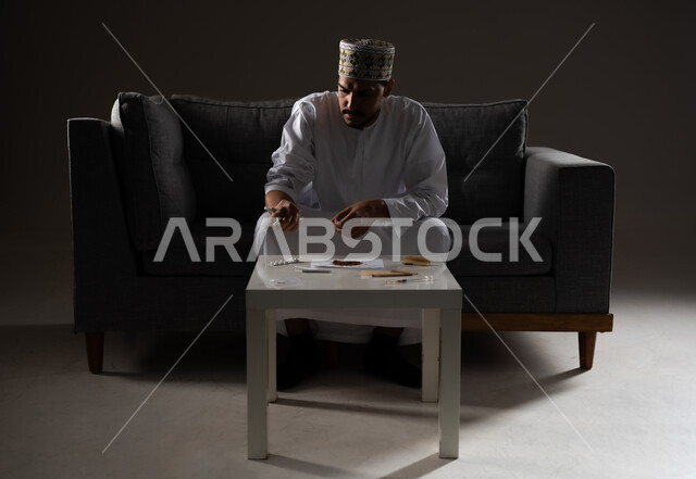 القيام بعادات سلوكية ضارة وتعاطي المواد السامة، الأثار السلبية للادمان على التبغ والمخدرات، المعاناة من الاضطرابات النفسية والمشاكل الصحية، بورتريه لرجل عربي خليجي عماني يرتدي الدشداشه والكمه يجلس على اريكة ويقوم بلف سجائر الحشيش المخدر، صوره ظليه