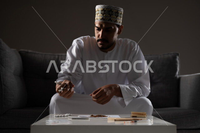 القيام بعادات سلوكية ضارة وتعاطي المواد السامة، الأثار السلبية للادمان على التبغ والمخدرات، المعاناة من الاضطرابات النفسية والمشاكل الصحية، بورتريه لرجل عربي خليجي عماني يرتدي الدشداشه والكمه يجلس على اريكة ويقوم بلف سجائر الحشيش المخدر، صوره ظليه