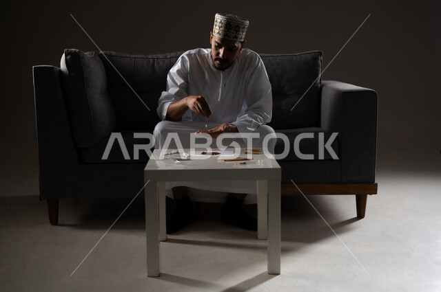 القيام بعادات سلوكية ضارة وتعاطي المواد السامة، الأثار السلبية للادمان على التبغ والمخدرات، المعاناة من الاضطرابات النفسية والمشاكل الصحية، بورتريه لرجل عربي خليجي عماني يرتدي الدشداشه والكمه يجلس على اريكة ويقوم بلف سجائر الحشيش المخدر، صوره ظليه