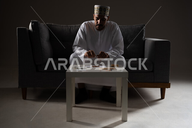 القيام بعادات سلوكية ضارة وتعاطي المواد السامة، الأثار السلبية للادمان على التبغ والمخدرات، المعاناة من الاضطرابات النفسية والمشاكل الصحية، بورتريه لرجل عربي خليجي عماني يرتدي الدشداشه والكمه يجلس على اريكة ويقوم بلف سجائر الحشيش المخدر، صوره ظليه