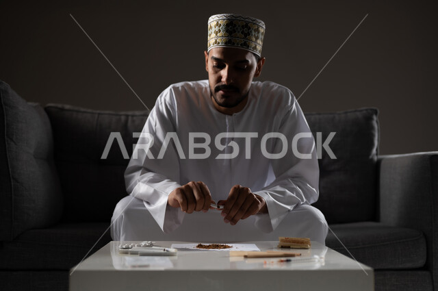 القيام بعادات سلوكية ضارة وتعاطي المواد السامة، الأثار السلبية للادمان على التبغ والمخدرات، المعاناة من الاضطرابات النفسية والمشاكل الصحية، بورتريه لرجل عربي خليجي عماني يرتدي الدشداشه والكمه يجلس على اريكة ويقوم بلف سجائر الحشيش المخدر، صوره ظليه