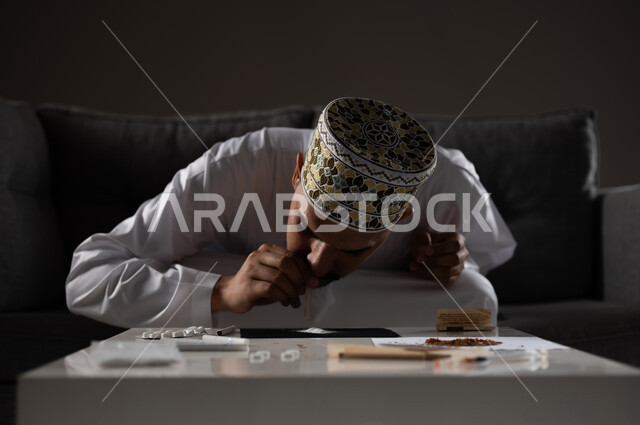 القيام بعادات سلوكية ضارة وتعاطي المواد السامة، الأثار السلبية للادمان على التبغ والمخدرات، المعاناة من الاضطرابات النفسية والمشاكل الصحية، بورتريه لرجل عربي خليجي عماني يرتدي الدشداشه والكمه يجلس على اريكة ويقوم باستنشاق المواد المخدره، صوره ظليه