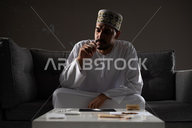 القيام بعادات سلوكية ضارة وتعاطي المواد السامة، الأثار السلبية للادمان على التبغ والمخدرات، المعاناة من الاضطرابات النفسية والمشاكل الصحية، بورتريه لرجل عربي خليجي عماني يرتدي الدشداشه والكمه يجلس على اريكة ويقوم بتدخين سجائر الحشيش المخدر، صوره ظليه