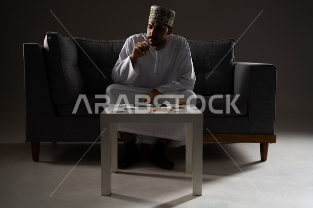 القيام بعادات سلوكية ضارة وتعاطي المواد السامة، الأثار السلبية للادمان على التبغ والمخدرات، المعاناة من الاضطرابات النفسية والمشاكل الصحية، بورتريه لرجل عربي خليجي عماني يرتدي الدشداشه والكمه يجلس على اريكة ويقوم بتدخين سجائر الحشيش المخدر، صوره ظليه