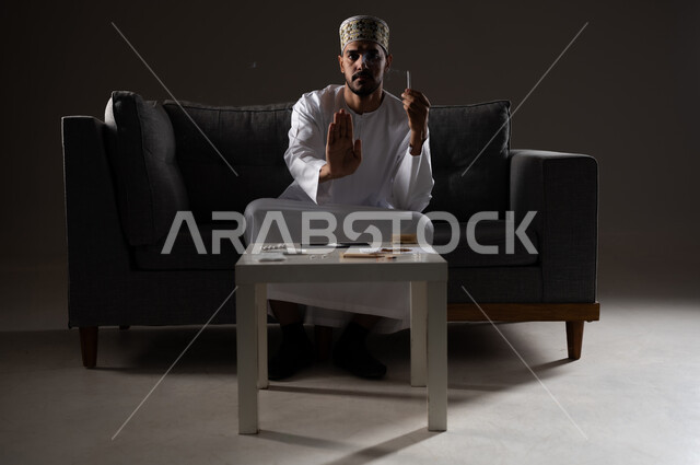 القيام بعادات سلوكية ضارة وتعاطي المواد السامة، الأثار السلبية للادمان على التبغ والمخدرات، المعاناة من الاضطرابات النفسية والمشاكل الصحية، بورتريه لرجل عربي خليجي عماني يرتدي الدشداشه والكمه يجلس على اريكة يمسك السيجاره بيده ويشير بكفه نحو الامام بايماءات التوقف عن التدخين، صوره ظليه