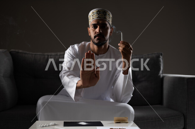 القيام بعادات سلوكية ضارة وتعاطي المواد السامة، الأثار السلبية للادمان على التبغ والمخدرات، المعاناة من الاضطرابات النفسية والمشاكل الصحية، بورتريه لرجل عربي خليجي عماني يرتدي الدشداشه والكمه يجلس على اريكة يمسك السيجاره بيده ويشير بكفه نحو الامام بايماءات التوقف عن التدخين، صوره ظليه