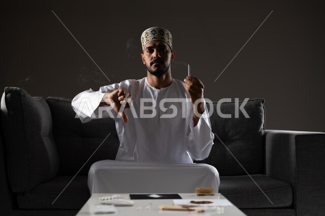 القيام بعادات سلوكية ضارة وتعاطي المواد السامة، الأثار السلبية للادمان على التبغ والمخدرات، المعاناة من الاضطرابات النفسية والمشاكل الصحية، بورتريه لرجل عربي خليجي عماني يرتدي الدشداشه والكمه يجلس على اريكة يمسك السيجاره بيده ويشير باصبع الابهام نحو الاسفل، صوره ظليه
