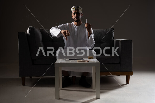 القيام بعادات سلوكية ضارة وتعاطي المواد السامة، الأثار السلبية للادمان على التبغ والمخدرات، المعاناة من الاضطرابات النفسية والمشاكل الصحية، بورتريه لرجل عربي خليجي عماني يرتدي الدشداشه والكمه يجلس على اريكة يمسك السيجاره بيده ويشير باصبع الابهام نحو الاسفل، صوره ظليه