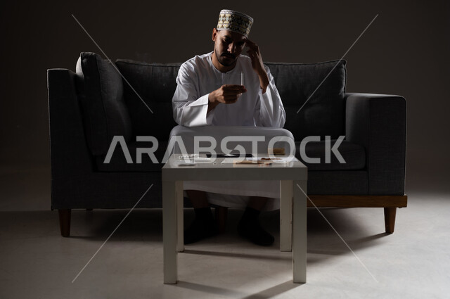 القيام بعادات سلوكية ضارة وتعاطي المواد السامة، الأثار السلبية للادمان على التبغ والمخدرات، المعاناة من الاضطرابات النفسية والمشاكل الصحية، بورتريه لرجل عربي خليجي عماني يرتدي الدشداشه والكمه يجلس على اريكة ويقوم بتدخين سجائر الحشيش المخدر، صوره ظليه