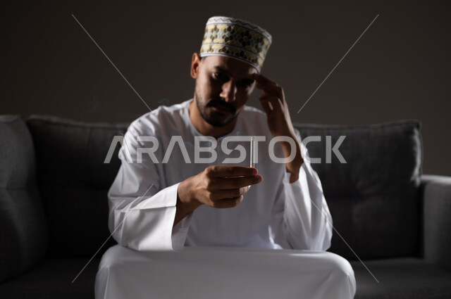 القيام بعادات سلوكية ضارة وتعاطي المواد السامة، الأثار السلبية للادمان على التبغ والمخدرات، المعاناة من الاضطرابات النفسية والمشاكل الصحية، بورتريه لرجل عربي خليجي عماني يرتدي الدشداشه والكمه يجلس على اريكة ويقوم بتدخين سجائر الحشيش المخدر، صوره ظليه