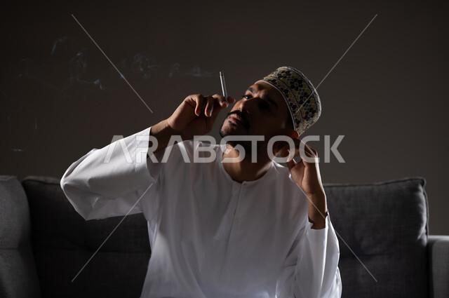 القيام بعادات سلوكية ضارة وتعاطي المواد السامة، الأثار السلبية للادمان على التبغ والمخدرات، المعاناة من الاضطرابات النفسية والمشاكل الصحية، بورتريه لرجل عربي خليجي عماني يرتدي الدشداشه والكمه يجلس على اريكة ويقوم بتدخين سجائر الحشيش المخدر، صوره ظليه