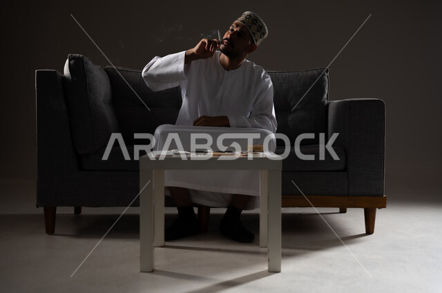 القيام بعادات سلوكية ضارة وتعاطي المواد السامة، الأثار السلبية للادمان على التبغ والمخدرات، المعاناة من الاضطرابات النفسية والمشاكل الصحية، بورتريه لرجل عربي خليجي عماني يرتدي الدشداشه والكمه يجلس على اريكة ويقوم بتدخين سجائر الحشيش المخدر، صوره ظليه