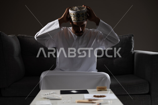معاقبة النفس وتأنيب الضمير، المعاناة من الاضطرابات النفسية والمشاكل الصحية، بورتريه لرجل عربي خليجي عماني يرتدي الدشداشه والكمه يجلس على الاريكة يقوم بالاستعداد لتناول المواد المخدره، الآثار السلبية للادمان والتعاطي، القيام بعادات سلوكية ضارة وتعاطي المواد السامة، صوره ظليه
