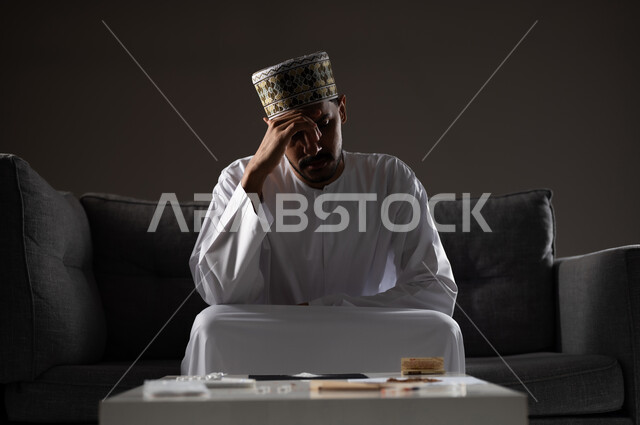 معاقبة النفس وتأنيب الضمير، المعاناة من الاضطرابات النفسية والمشاكل الصحية، بورتريه لرجل عربي خليجي عماني يرتدي الدشداشه والكمه يجلس على الاريكة بتعابير التعب والندم، الآثار السلبية للادمان والتعاطي، القيام بعادات سلوكية ضارة وتعاطي المواد السامة، صوره ظليه
