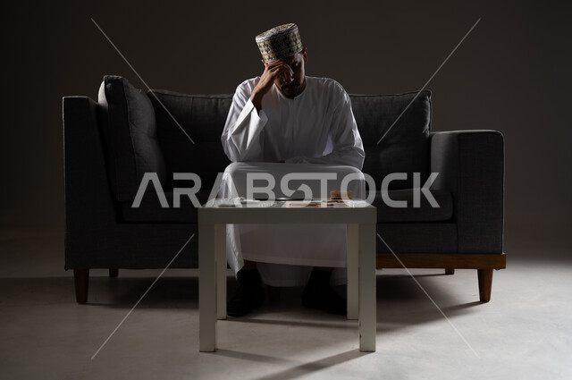 معاقبة النفس وتأنيب الضمير، المعاناة من الاضطرابات النفسية والمشاكل الصحية، بورتريه لرجل عربي خليجي عماني يرتدي الدشداشه والكمه يجلس على الاريكة بتعابير التعب والندم، الآثار السلبية للادمان والتعاطي، القيام بعادات سلوكية ضارة وتعاطي المواد السامة، صوره ظليه