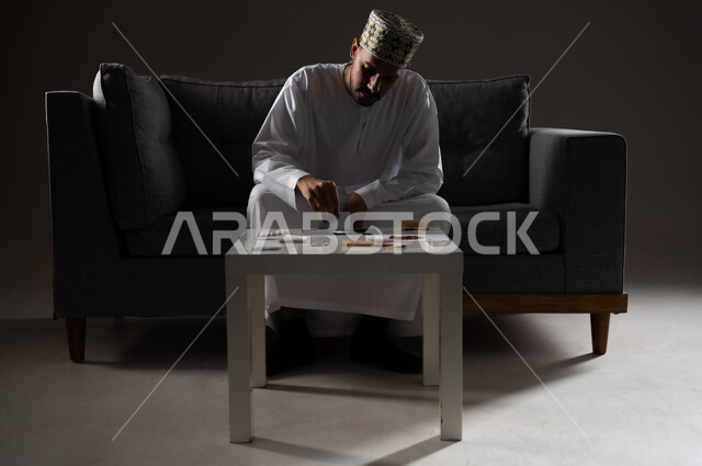 القيام بعادات سلوكية ضارة وتعاطي المواد السامة، الأثار السلبية للادمان على التبغ والمخدرات، المعاناة من الاضطرابات النفسية والمشاكل الصحية، بورتريه لرجل عربي خليجي عماني يرتدي الدشداشه والكمه يجلس على اريكة ويقوم بتحضير المواد المخدره استعدادا لتعاطيها، صوره ظليه