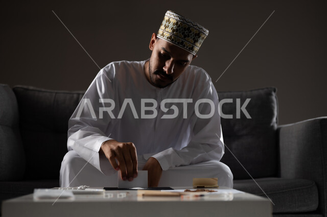 القيام بعادات سلوكية ضارة وتعاطي المواد السامة، الأثار السلبية للادمان على التبغ والمخدرات، المعاناة من الاضطرابات النفسية والمشاكل الصحية، بورتريه لرجل عربي خليجي عماني يرتدي الدشداشه والكمه يجلس على اريكة ويقوم بتحضير المواد المخدره استعدادا لتعاطيها، صوره ظليه