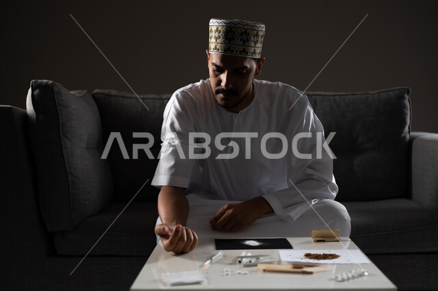 القيام بعادات سلوكية ضارة وتعاطي المواد السامة، الأثار السلبية للادمان على التبغ والمخدرات، المعاناة من الاضطرابات النفسية والمشاكل الصحية، بورتريه لرجل عربي خليجي عماني يرتدي الدشداشه والكمه يجلس على اريكة وييكشف عن ذراعه استعدادا لحقن المواد المخدره، صوره ظليه