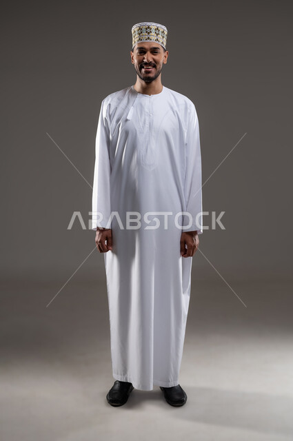 التقاط الصور الشخصية للمناسبات العامه، الاهتمام بالأناقة والمظهر الخارجي, مفهوم الرجولة والثقة بالنفس، بورتريه لرجل عربي خليجي عماني مبتسم يرتدي الدشداشة و الكمه ينظر الى الكاميرا بإيماءات السرور والسعادة، صورة الجسم بالطول الكامل, خلفية بيضاء