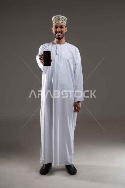 Using advanced modern technology and techniques, portrait of a smiling young Arab Gulf Omani man wearing a dishdasha and a sleeve displaying a blank black screen via a mobile phone, looking at the camera with an expression of pleasure, full-length body image, white background