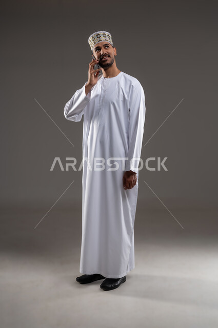 تبادل الأحاديث مع الأهل والأصدقاء، إجراء مكالمة هاتفية باستخدام الهاتف المحمول، بورتريه بالطول الكامل لرجل عربي خليجي عماني يرتدي الدشداشة والكمه يقوم بالتحدث بهاتفهه المحمول بتعابير الفرح والسرور، خلفية بيضاء