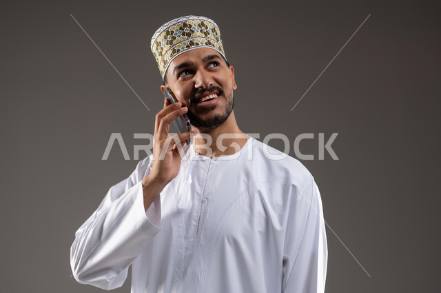 تبادل الأحاديث مع الأهل والأصدقاء، إجراء مكالمة هاتفية باستخدام الهاتف المحمول، بورتريه مقرب لرجل عربي خليجي عماني يرتدي الدشداشة والكمه يقوم بالتحدث بهاتفهه المحمول بتعابير الفرح والسرور، خلفية بيضاء