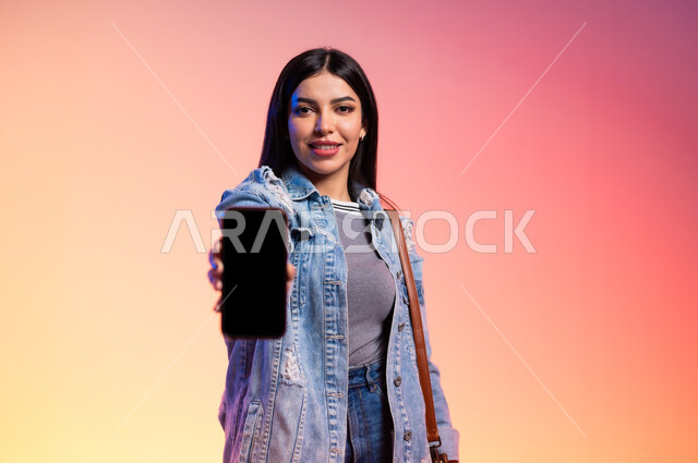 ملامح الانوثة والنعومة, ألبسة خليجية نسائية عصرية، بورتريه مقرب لامرأة خليجية سعودية ترتدي اللباس الكاجوال, تقوم بعرض شاشة سوداء فارغة، مفهوم الموضة والأناقة، عرض الأزياء والموديلات الحديثة، شراءترتدي اللباس الكاجوا الملابس والاكسسوارات الجديدة، خلفية ملونة