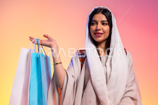 الاستمتاع بالتسوق وأثره على نفسية النساء، عرض الأزياء والموديلات الحديثة، ألبسة خليجية نسائية عصرية، بورتريه لامرأة عربية خليجية سعودية ترتدي عباءة باللون البيج تحمل بيدها أكياس ورقية ملونة، مفهوم الموضة والأناقة، شراء الملابس والاكسسوارات الجديدة، خلفية ملونة