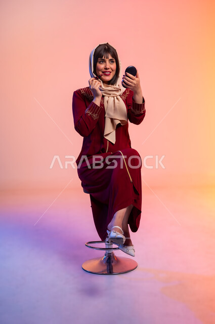 الاهتمام بالانوثة والمظهر الخارجي, ألبسة خليجية نسائية عصرية، بورتريه لامرأة عربية خليجية سعودية ترتدي العباءة تقوم بوضع احد مساحيق التجميل، مفهوم الموضة والأناقة، عرض الأزياء والموديلات الحديثة، شراء الملابس والاكسسوارات الجديدة، خلفية ملونة