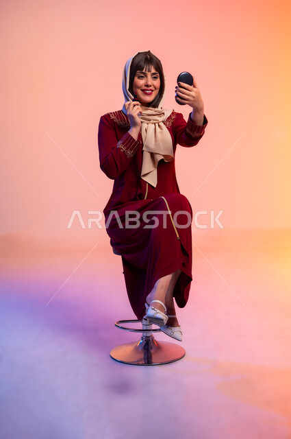 الاهتمام بالانوثة والمظهر الخارجي, ألبسة خليجية نسائية عصرية، بورتريه لامرأة عربية خليجية سعودية ترتدي العباءة تقوم بوضع احد مساحيق التجميل، مفهوم الموضة والأناقة، عرض الأزياء والموديلات الحديثة، شراء الملابس والاكسسوارات الجديدة، خلفية ملونة