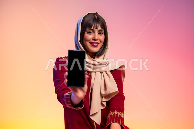 عرض الأزياء والموديلات الحديثة، شراء الملابس الجديدة، ألبسة خليجية نسائية عصرية، بورتريه لامرأة عربية خليجية سعودية ترتدي عباءة باللون الخمري تحمل الهاتف المحمول تقوم بعرض شاشة سوداء فارغة، مفهوم الموضة والأناقة، خلفية ملونة