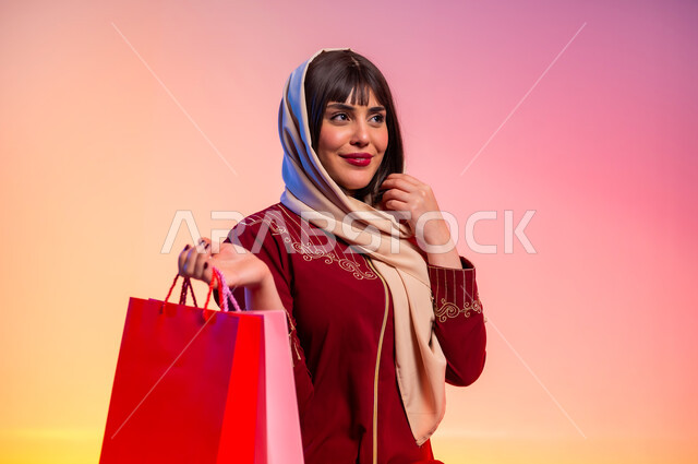عرض الأزياء والموديلات الحديثة، شراء الملابس الجديدة، أثر التسوق على نفسية النساء، ألبسة خليجية نسائية عصرية، بورتريه لامرأة عربية خليجية سعودية ترتدي عباءة باللون الخمري تحمل أكياس ورقية ملونة، مفهوم الموضة والأناقة، خلفية ملونة