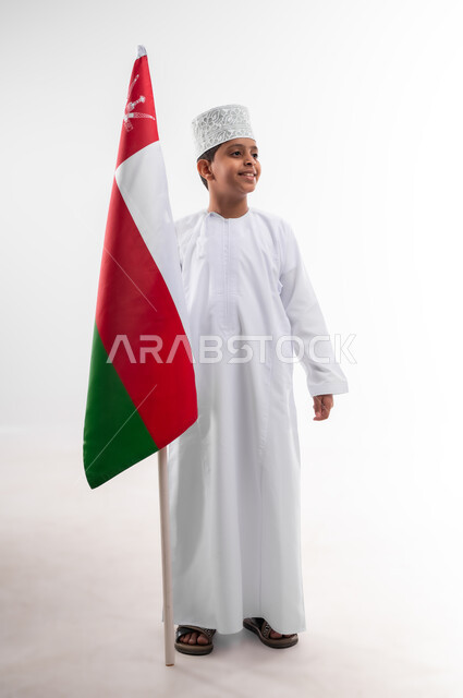 إحياء ذكرى اليوم الوطني العماني، الرايات والأعلام الوطنية للدول العربية، الاعتزاز والفخر بالرموز والراية العمانية، بورتريه لطفل عربي خليجي عماني يرتدي الدشداشة والكمه، مفهوم حب الوطن والإنتماء إليه، الجسم بالطول الكامل، خلفية بيضاء