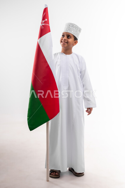 إحياء ذكرى اليوم الوطني العماني، الرايات والأعلام الوطنية للدول العربية، الاعتزاز والفخر بالرموز والراية العمانية، بورتريه لطفل عربي خليجي عماني يرتدي الدشداشة والكمه، مفهوم حب الوطن والإنتماء إليه، الجسم بالطول الكامل، خلفية بيضاء