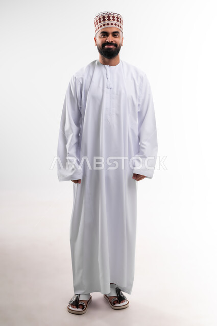 وقفة بايماءات ايدي متقاطعة بشكل مستقيم تدل على الثقة بالنفس، بورتريه لشاب عربي خليجي عماني يرتدي الدشداشة والكمه تظهر عليه تعابير السعادة والفرح، الإهتمام بالأناقة والمظهر الخارجي، الجسم بالطول الكامل، خلفية بيضاء
