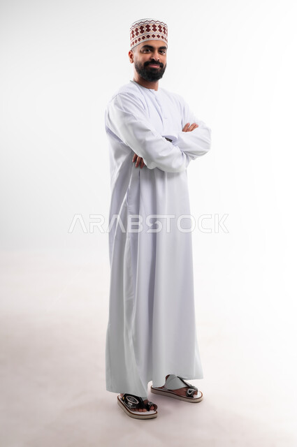 وقفة بايماءات ايدي متقاطعة بشكل مستقيم تدل على الثقة بالنفس، بورتريه لشاب عربي خليجي عماني يرتدي الدشداشة والكمه تظهر عليه تعابير السعادة والفرح، الإهتمام بالأناقة والمظهر الخارجي، الجسم بالطول الكامل، خلفية بيضاء