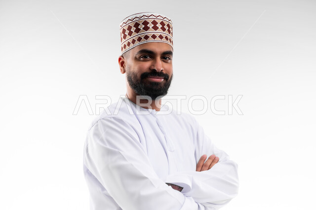 وقفة بايماءات ايدي متقاطعة بشكل مستقيم تدل على الثقة بالنفس، بورتريه لشاب عربي خليجي عماني يرتدي الدشداشة والكمه تظهر عليه تعابير السعادة والفرح، الإهتمام بالأناقة والمظهر الخارجي، الجسم بالطول الكامل، خلفية بيضاء