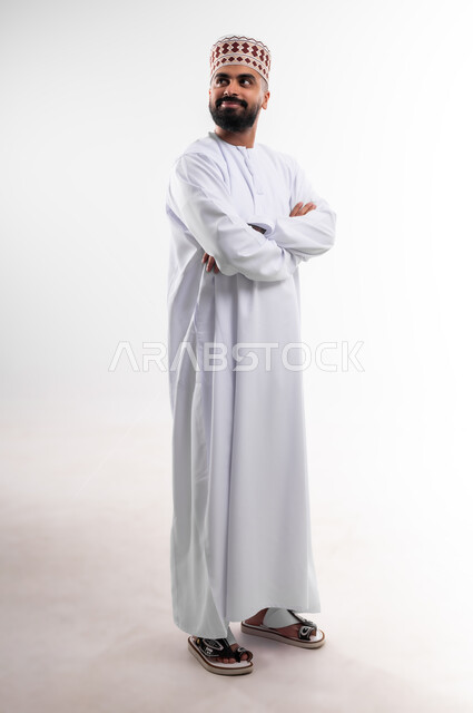 وقفة بايماءات ايدي متقاطعة بشكل مستقيم تدل على الثقة بالنفس، بورتريه لشاب عربي خليجي عماني يرتدي الدشداشة والكمه تظهر عليه تعابير السعادة والفرح، الإهتمام بالأناقة والمظهر الخارجي، الجسم بالطول الكامل، خلفية بيضاء