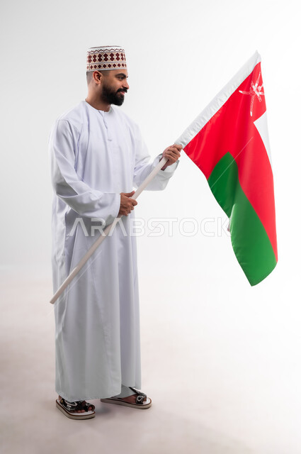 المشاركة بالاعياد والمناسبات الوطنية، الاحتفال بذكرى اليوم الوطني العماني، الاعتزاز والفخر بالرموز والراية العمانية، بورتريه لشاب عربي خليجي عماني يرتدي الدشداشة والكمه، مفهوم حب الوطن والإنتماء إليه، الجسم بالطول الكامل، خلفية بيضاء