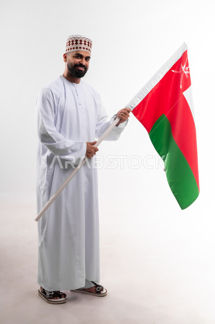 Participation in national holidays and occasions, celebration of Omani National Day, pride in Omani symbols and flag, portrait of a young Gulf Arab Omani man wearing a dishdasha and a kuma holding the flag of the Sultanate of Oman in his hand, the concept of love of the homeland and belonging to it, full body, white background