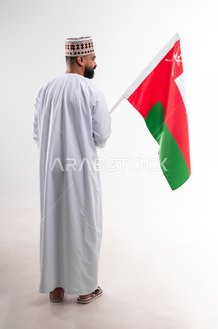 Participation in national holidays and occasions, celebration of Omani National Day, pride in Omani symbols and flag, portrait of a young Gulf Arab Omani man wearing a dishdasha and a kuma, concept of love of the homeland and belonging to it, full body, white background