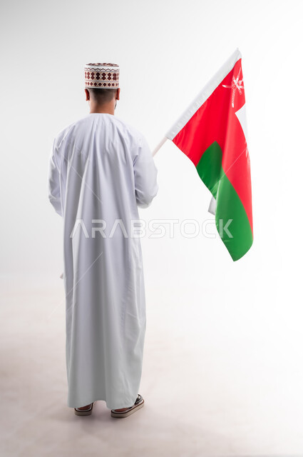 المشاركة بالاعياد والمناسبات الوطنية، الاحتفال بذكرى اليوم الوطني العماني، الاعتزاز والفخر بالرموز والراية العمانية، بورتريه لشاب عربي خليجي عماني يرتدي الدشداشة والكمه، مفهوم حب الوطن والإنتماء إليه، الجسم بالطول الكامل، خلفية بيضاء