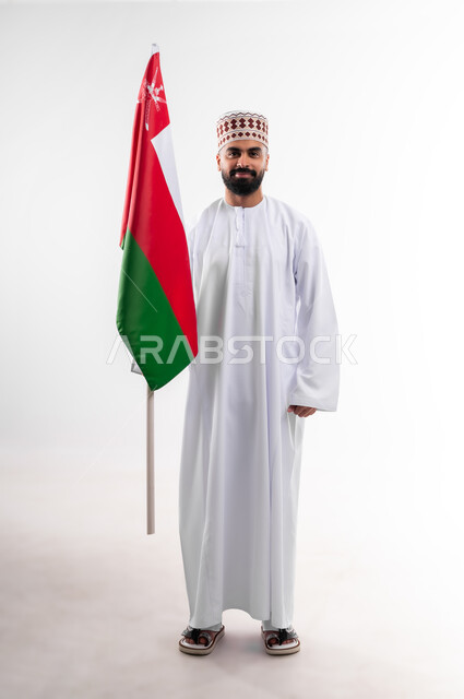 Participation in national holidays and occasions, celebration of Omani National Day, pride in Omani symbols and flag, portrait of a young Gulf Arab Omani man wearing a dishdasha and a kuma holding the flag of the Sultanate of Oman in his hand, the concept of love of the homeland and belonging to it, full body, white background