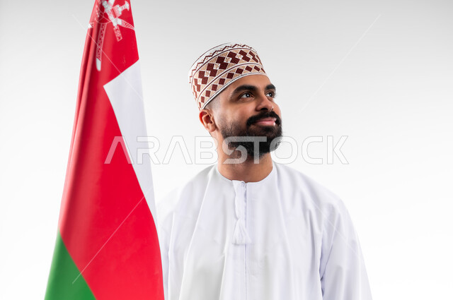 الفخر والاعتزاز بالانتماء للهوية، المشاركة بالاعياد والمناسبات الوطنية، الاحتفال بذكرى اليوم الوطني العماني، الاعتزاز والفخر بالرموز والراية العمانية، بورتريه لشاب عربي خليجي عماني يرتدي الدشداشة والكمه، مفهوم حب الوطن، خلفية بيضاء