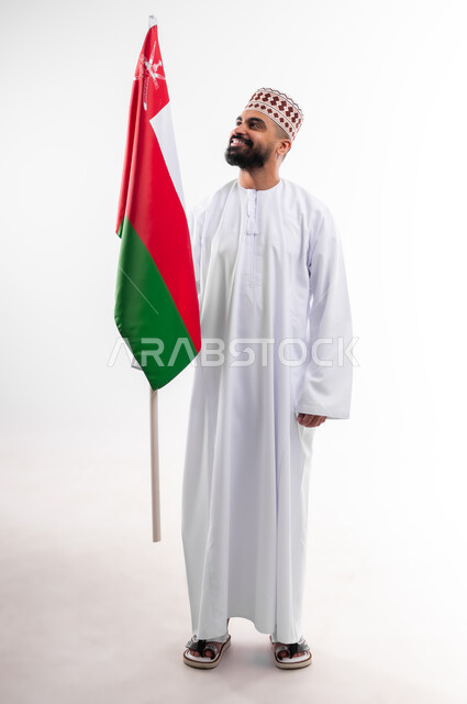 المشاركة بالاعياد والمناسبات الوطنية، الاحتفال بذكرى اليوم الوطني العماني، الاعتزاز والفخر بالرموز والراية العمانية، بورتريه لشاب عربي خليجي عماني يرتدي الدشداشة والكمه، مفهوم حب الوطن والإنتماء إليه، الجسم بالطول الكامل، خلفية بيضاء