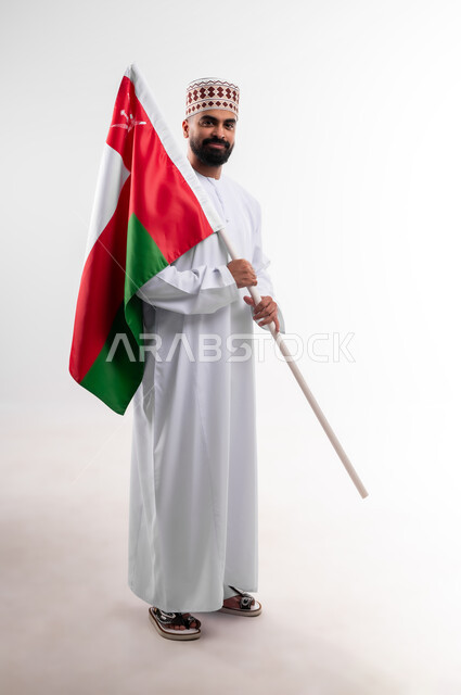 Participation in national holidays and occasions, celebration of Omani National Day, pride in Omani symbols and flag, portrait of a young Gulf Arab Omani man wearing a dishdasha and a kuma holding the flag of the Sultanate of Oman in his hand, the concept of love of the homeland and belonging to it, full body, white background